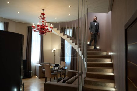 Photo for Middle-aged man goes down the stairs to the living room, indoors designer chandelier - Royalty Free Image