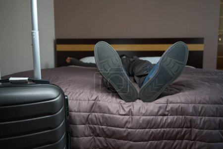 Téléchargez les photos : Voyageur fatigué se repose de la route dans les chaussures sur un grand lit dans la chambre d'hôtel, à côté de la valise de voyage - en image libre de droit