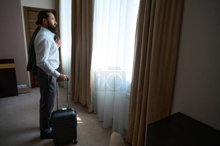 Foto de Viajando hombre de mediana edad se para en la ventana de una habitación de hotel, tiene una maleta de viaje - Imagen libre de derechos
