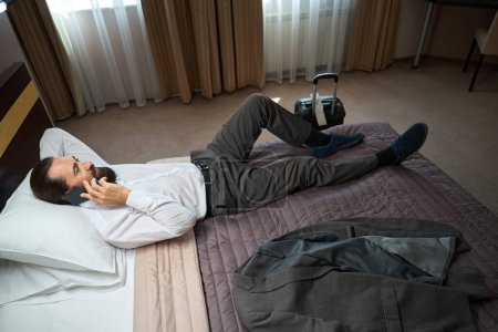 Foto de Hombre con el teléfono tumbado para descansar de la carretera, junto a la chaqueta de viaje y la maleta - Imagen libre de derechos