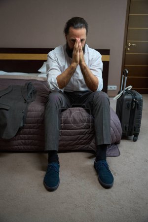 Téléchargez les photos : Homme assis pour se reposer sur le bord du lit dans la chambre d'hôtel, il était fatigué de longue route - en image libre de droit