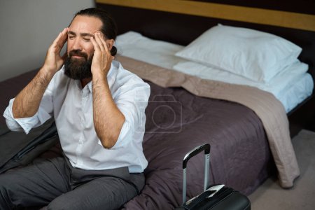 Foto de Viajero con dolor de cabeza se sentó a descansar en una cama en una habitación de hotel, estaba cansado de la carretera - Imagen libre de derechos