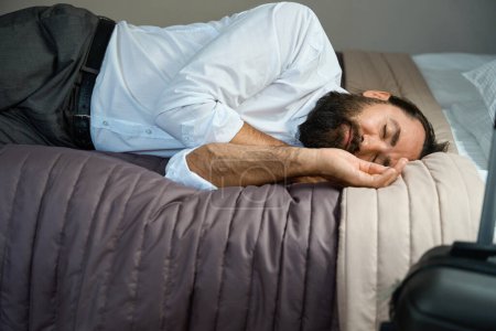 Foto de Varón cansado con dolor de cabeza se acostó a descansar en la cama en una habitación de hotel, junto a la maleta de viaje - Imagen libre de derechos