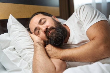Téléchargez les photos : Homme barbu dort dans une chambre d'hôtel lumineuse, la chambre est propre et confortable - en image libre de droit
