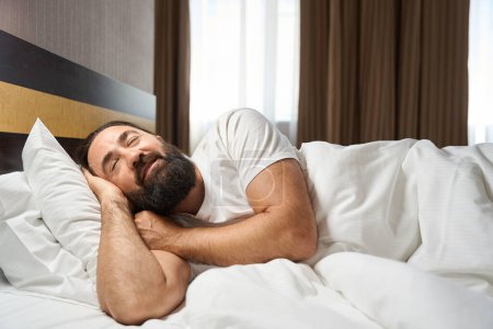 Téléchargez les photos : Homme d'âge moyen dort sur le côté sur un grand lit, la chambre est propre et confortable - en image libre de droit