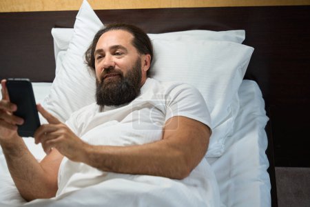 Téléchargez les photos : Homme adulte lit les nouvelles dans un téléphone portable, il se trouve sur un grand lit - en image libre de droit