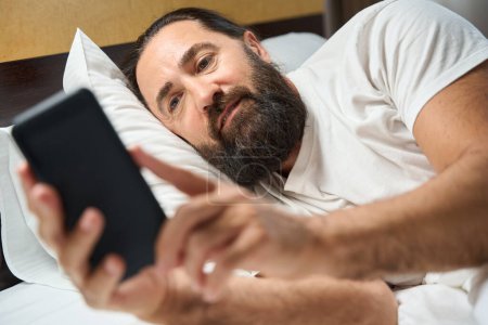 Foto de Hombre despierto lee las noticias en un teléfono móvil, yace en una cama grande - Imagen libre de derechos