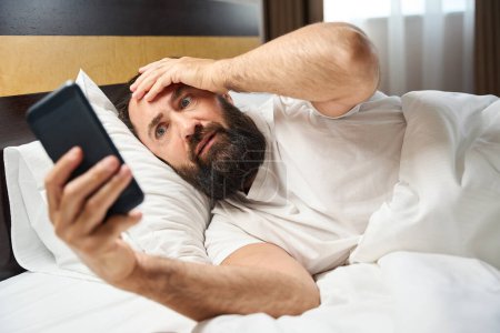 Foto de El hombre se acuesta en la cama con un teléfono móvil, tiene una barba gruesa bien arreglada - Imagen libre de derechos