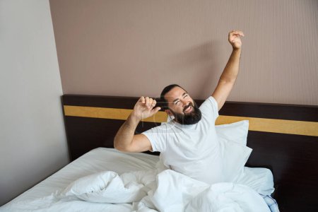 Téléchargez les photos : Homme joyeux se réveille dans une chambre d'hôtel lumineuse, la chambre est propre et confortable - en image libre de droit