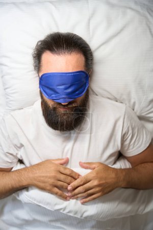 Téléchargez les photos : Homme dort avec un bandeau sur un lit blanc neige, il a une barbe épaisse bien entretenue - en image libre de droit