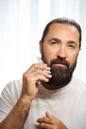 Photo for Adult male combing his thick beard with a special comb, a man in white clothes - Royalty Free Image
