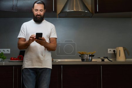 Foto de Hombre barbudo se para en el área de la cocina y mensajes de texto en un teléfono móvil, él está en pantalones vaqueros cómodos - Imagen libre de derechos