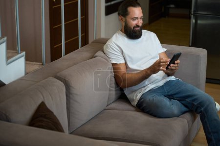 Foto de Sonriente hombre pasa la noche charlando en línea en el teléfono móvil, él está usando pantalones vaqueros cómodos - Imagen libre de derechos