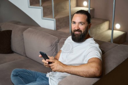 Téléchargez les photos : Beau homme passe la soirée à bavarder en ligne sur un téléphone portable, il est en jean confortable - en image libre de droit