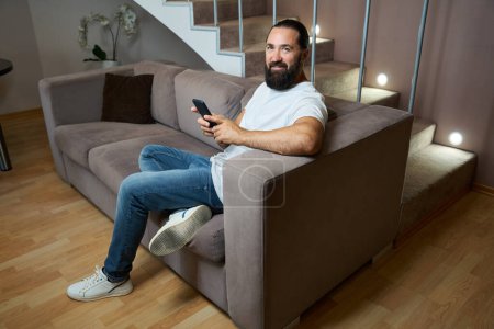 Photo for Hotel guest spends the evening online chatting on a mobile phone, he is in comfortable casual clothes - Royalty Free Image