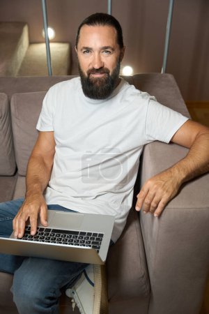 Téléchargez les photos : Beau homme assis sur un canapé avec un ordinateur portable, il est dans des vêtements décontractés confortables - en image libre de droit