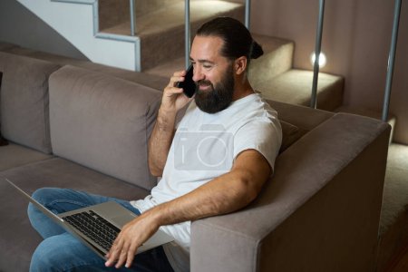Téléchargez les photos : Homme souriant communique sur un téléphone portable et travaille sur un ordinateur portable moderne, il est dans des vêtements décontractés confortables - en image libre de droit