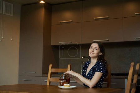 Téléchargez les photos : Jolie femme dans une robe à pois boit du thé avec un gâteau, elle est assise dans la cuisine à la table - en image libre de droit