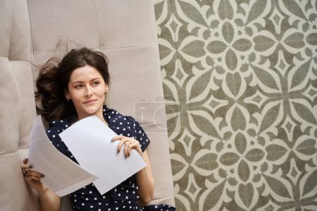 Téléchargez les photos : Femme rêveuse dans une robe à pois étudie les papiers de travail, elle est assise sur un canapé doux - en image libre de droit