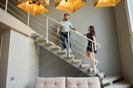 Téléchargez les photos : Mari et femme montent les escaliers dans la maison, ils se tiennent tendrement la main - en image libre de droit