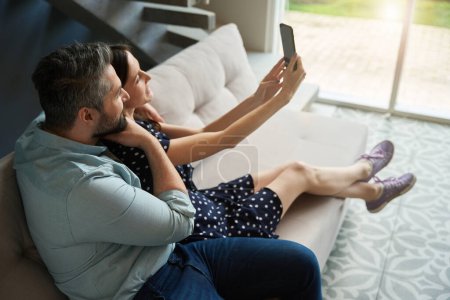 Photo for Middle-aged couple rest comfortably on a soft sofa, they take a selfie - Royalty Free Image