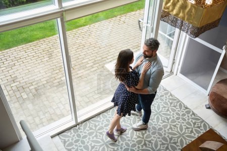 Foto de Marido feliz y la esposa están de pie abrazando en una amplia sala de estar, fuera de la ventana es un lindo patio trasero - Imagen libre de derechos