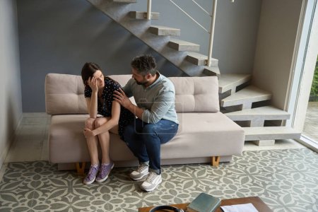 Foto de Hombre adulto abraza y calma a una mujer encantadora en la sala de estar, la pareja se encuentra en un sofá ligero - Imagen libre de derechos