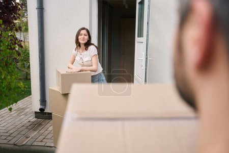 Téléchargez les photos : Jeune femme appuyée sur des boîtes et regardant la caméra tout en se tenant à l'entrée de la maison - en image libre de droit