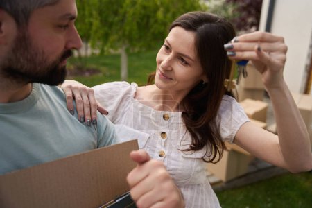Photo for Pretty female putting hand on shoulder of man and showing keys to him while he holding box in yard - Royalty Free Image