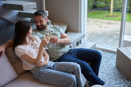 Téléchargez les photos : Femme et homme caucasiens assis sur le canapé tout en se regardant et cliquetis verres à vin à l'intérieur - en image libre de droit