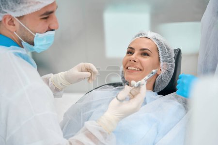 Foto de Mujer joven se sienta en una silla dental, el médico sostiene una jeringa con analgésicos en las manos - Imagen libre de derechos