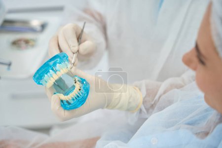 Foto de El médico ortodoncista le muestra al paciente un modelo con implantes dentales y carillas, un médico con guantes protectores - Imagen libre de derechos