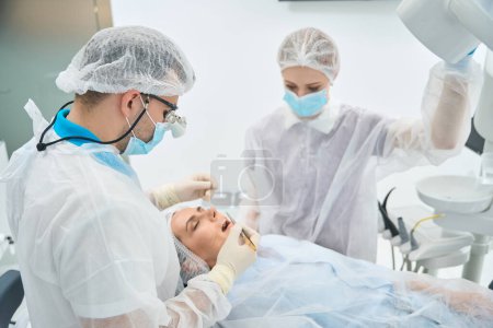 Téléchargez les photos : Un dentiste masculin traite un canal dentaire à une jeune femme au microscope, à proximité d'un assistant en uniforme médical - en image libre de droit