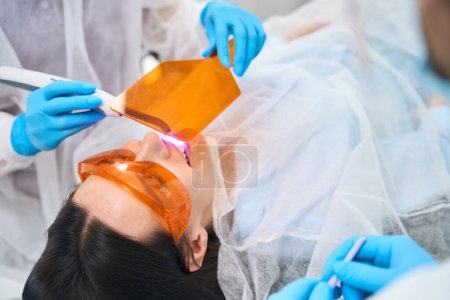 Foto de Dentista con la ayuda de un asistente llena el diente de los pacientes, el asistente utiliza una pantalla protectora - Imagen libre de derechos
