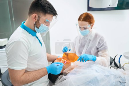 Foto de Médico masculino pone un relleno de fotopolímero en un paciente, un asistente utiliza una pantalla protectora - Imagen libre de derechos