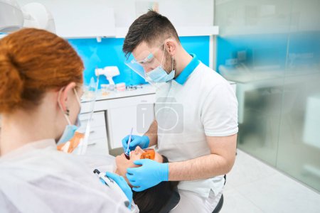 Foto de Médico en un consultorio dental pone un relleno de fotopolímero en un paciente, un asistente de pelo rojo está cerca - Imagen libre de derechos