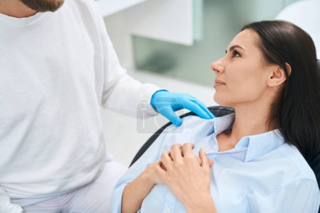 Foto de Dentista masculino en uniforme blanco y guantes protectores calmando a paciente mujer nerviosa antes del chequeo de salud de la cavidad oral en asiento cómodo - Imagen libre de derechos