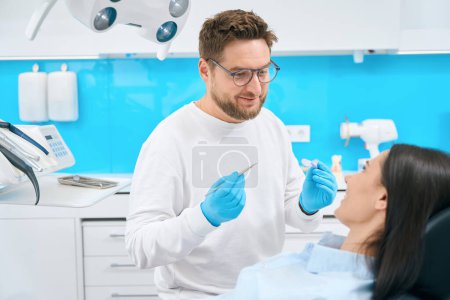 Téléchargez les photos : Médecin sympathique stomatologue tenant miroir buccal et sonde de faucille pour trouver la carie dentaire ou d'autres problèmes buccodentaires potentiels chez la patiente, check-up - en image libre de droit