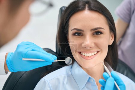 Foto de Mujer sonriente sentada en silla de ortodoncia y esperando dentista calificado examinando sus dientes y encías con un espejo bucal especial y sonda de hoz - Imagen libre de derechos