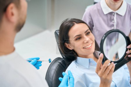Foto de Mujer hermosa paciente de clínica estomatológica privada mirando el espejo en sus dientes después de la limpieza profesional y blanqueamiento, procedimiento de rutina - Imagen libre de derechos