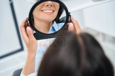 Foto de Odontóloga mujer cercana mirando el espejo a sus dientes y sonriendo, satisfecha con dientes sanos y blancos y aliento fresco - Imagen libre de derechos