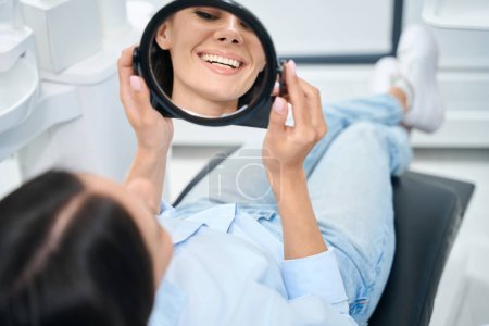 Foto de Mujer atractiva paciente acostada en la silla de ortodoncia y mirando al espejo en su nueva sonrisa sana y blanca, dentadura postiza o instalación de puente, servicio de clínica dental - Imagen libre de derechos