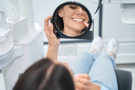Foto de Mujer alegre mirando al espejo y disfrutando de su nueva prótesis dental, sonriendo y tocando nuevos dientes, acostada en silla de ortodoncia en la consulta del dentista - Imagen libre de derechos
