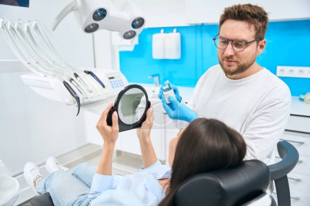 Téléchargez les photos : Technicien dentaire barbu suggérant quelques prothèses dentaires ou vésicules à la cliente qui se trouve dans sa chaise dentaire, clinique privée servise - en image libre de droit