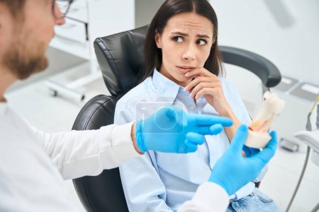 Foto de Mujer asustada paciente escuchando atentamente al técnico dental que explica la forma de su tratamiento dental en el modelo de dientes de plástico, clínica privada - Imagen libre de derechos
