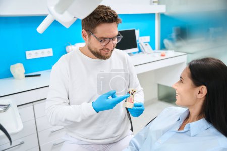 Téléchargez les photos : Dentiste souriant en uniforme tenant le modèle 3D des dents et expliquant à la patiente comment il traitera le canal radiculaire - en image libre de droit