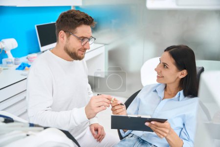 Photo for Professional stomatologist acquainting female patient with course of treatment and offering to sign insurance documents and treatment agreement - Royalty Free Image