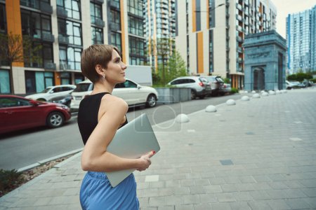Foto de Mujer joven camina por una calle de la ciudad con un ordenador portátil, alrededor de grandes edificios modernos - Imagen libre de derechos
