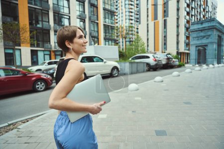 Foto de Mujer delgada camina a lo largo de una calle de la ciudad con un ordenador portátil, alrededor de grandes edificios modernos - Imagen libre de derechos