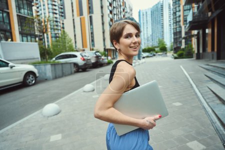 Photo for Beautiful young female walks along a city street with a laptop, she is in comfortable youth clothes - Royalty Free Image